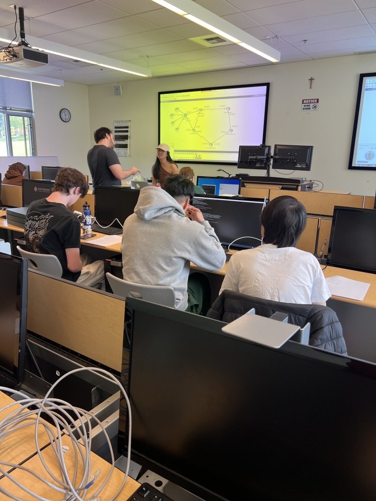 Students sit work at rows of computers, with a diagram on a screen at front of classroom