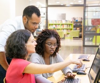 An instructor leans down to help two students working at a computer.