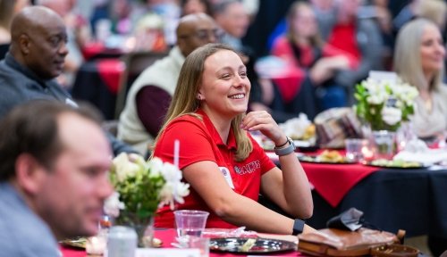 A crowd of alumni sit at round table with red tablecloths and white flower centerpieces, focus on a young woman in Saints red gear.