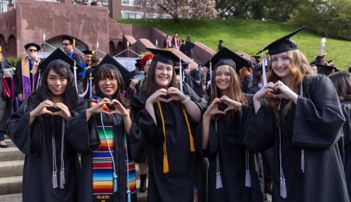 Five students in commencement regalia stand in a line, making hearts with their hands for the camera, the grand staircase in the background
