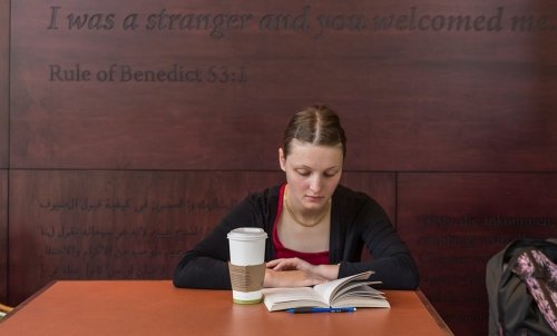 A student studies at a table with coffee and book in Harned Hall, with the quote "I was a stranger and you welcomed me" from Rule of Benedict 53:1 on the wall behind her.