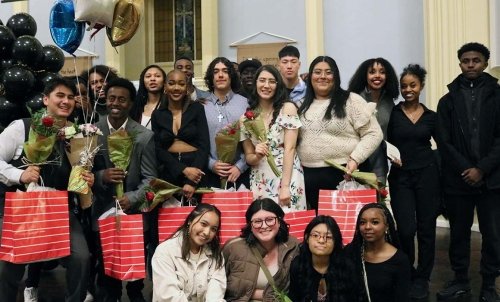 The fifth Act Six cadre poses, a group of 19 students holding red bags and balloons in the background