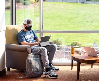 A masked student sits in a lounge area working on a laptop in front of a large window showing the grass outside.