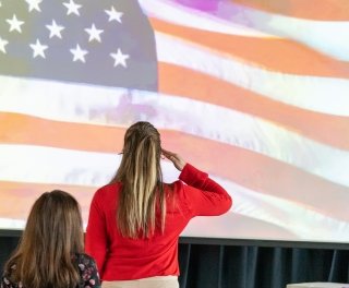 A woman in red stands and salutes the American flag projected on a screen.