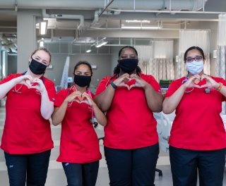 Four nursing students in scrubs and face masks pose making the Saints Care heart symbol with their hands.