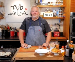 Celebrity chef Tom Douglas stands in his show kitchen, with lots of kitchen accoutrements around him, and the brand label "rub with love" behind him.