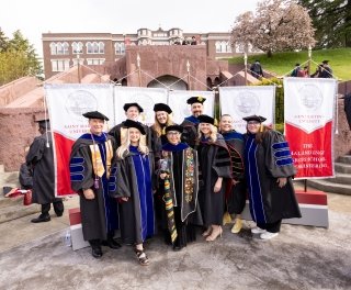 Ph.D. candidates pose in regalia at the food of the Saint Martin's grand staircase, with the college banners behind them and other graduates gathering around