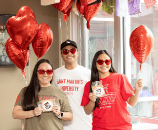 Three students in Saints shirts and wearing red heart sunglasses hold red heart balloons and 3.21GIVE cookies