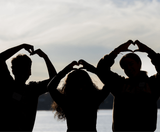 Three silhouettes of students making hearts with their hands in front of a twilit lake