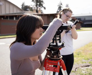 A students works with survey equipment while another takes notes outside