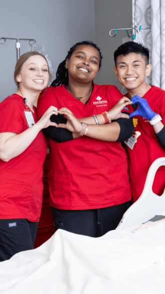 Three nursing students in red scrubs interlock their hands to make hearts