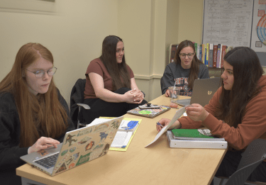 Four students sit around a table, tutors assisting students with work on their laptops