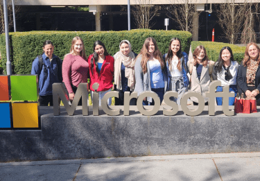 A group of students stand behind the Microsoft logo on the Microsoft campus