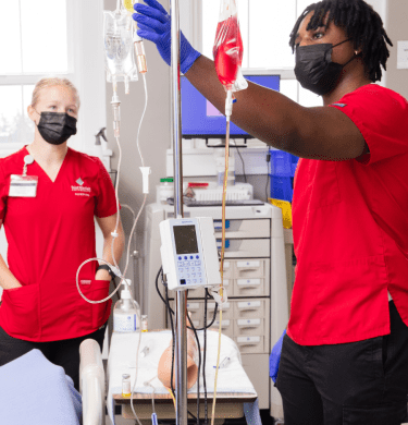 Two students in Saints red scrubs work with equipment in the nursing lab