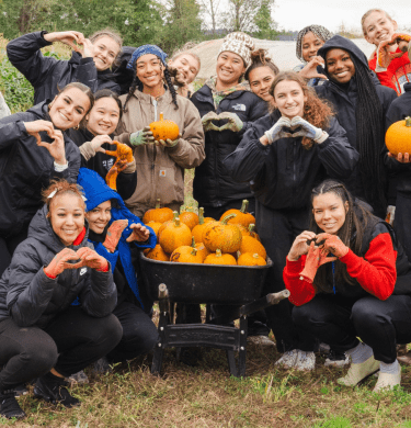 Group of students outside around a wheelbarrow full of pumpkins, making hearts with hands