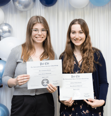 Two students stand with honor society certificates below a balloon arch