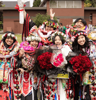 Students in graduation gowns and bedecked in flowers celebrate outside
