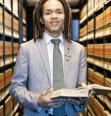 A student in a suit holds an open book surrounded by shelves filled with books