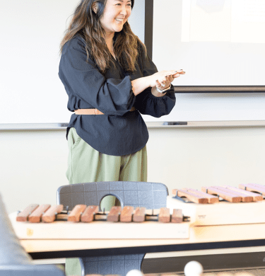 A student presents at the front of class, wooden xylophones on the desks