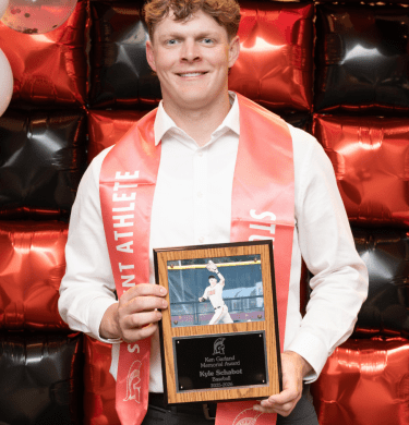 A student holds a baseball placard, wearing a red student-athlete sash in front of a balloon wall