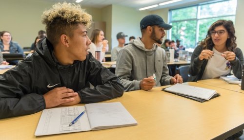 Three students seated in a row at a desk discuss with papers in front of them and a classroom full of other students behind.