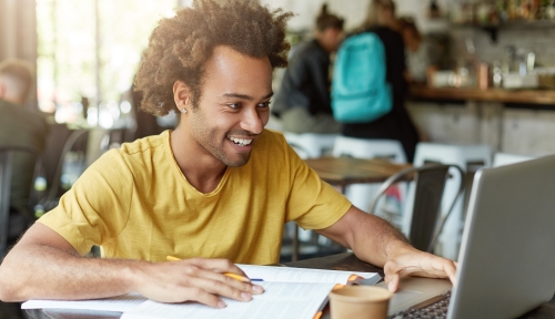 A student smiles, working at a laptop with an open notebook in a cafe.