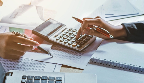 Close up of a hand holding a pencil working on a calculator with a laptop, lots, of paper, and another hand with a pen around it.