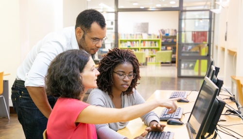 An instructor leans down to help two students working at a computer.