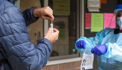 Someone puts a swab from a COVID test in a vial, while another person in PPE holds open a bag for them to put it in.