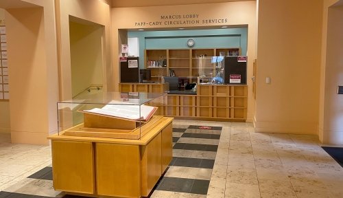 The entrance hall of the library, with the circulation desk in the background and a book in a glass cover in the foreground.
