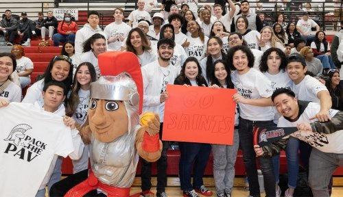 Students in white Pack the Pav tshirts pose in the bleachers, with Marty the mascot and President Bonds-Raacke in the front, holding a red sign that says GO SAINTS