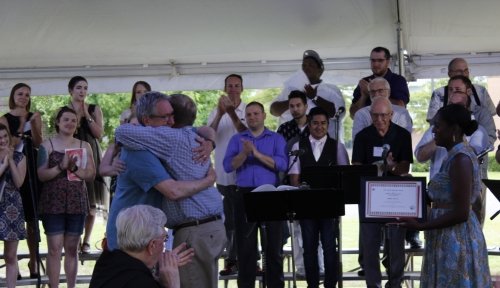 Two alumni hug in the foreground while a choir on risers clap in the background