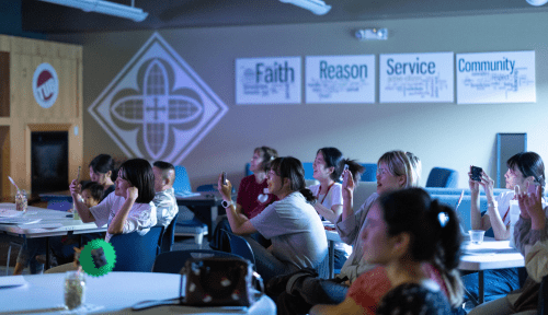 Students sitting at round tables hold up their phones toward a stage