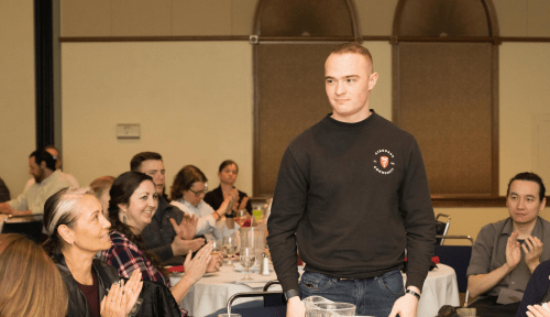 A veteran stands up from a table while a seated audience applauds him.