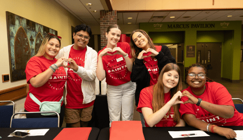 Students and staff wearing red Orientation shirts make hearts with hands behind registration desk