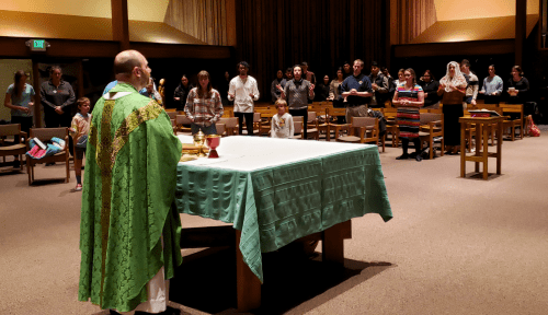 A Catholic father dressed in green vestments, stands in front of a large table, facing a crowd of students standing amongst pews