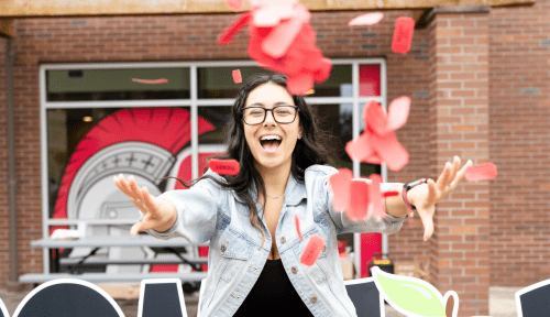 A student smiles, red tickets having just been thrown in the air drifting down in front of her, the Saints helmet on a window in the background