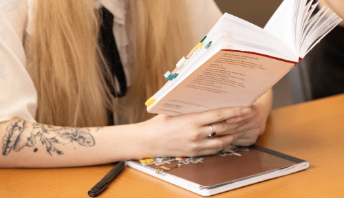 An arm with a floral tattoo holds an Oxford edition Shakespeare book with colored tabs, arms resting on a brown notebook with a pen nearby