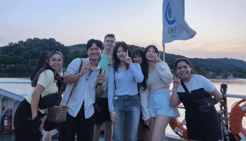 A group of students pose in front of water, the hills of Korea behind