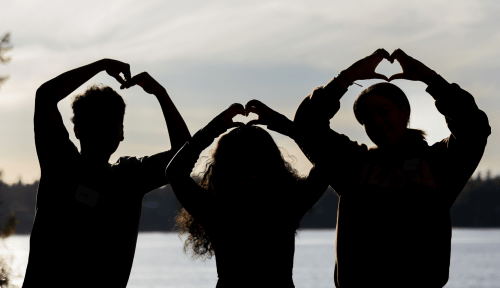 Three silhouettes of students making hearts with their hands in front of a twilit lake