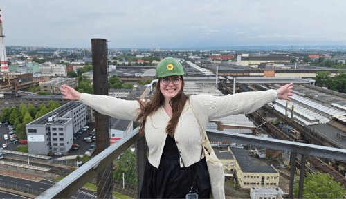 A student in a hard hat stands on top of building arms outstretched, the city laid out below her