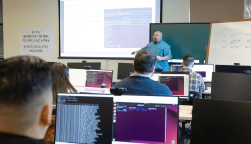 A teacher demonstrates at the front of class while students sit at computers