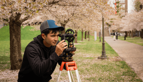 A student uses survey equipment outside beneath blossoming trees