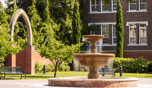Fountain in a courtyard with brick buildings and an arch behind it