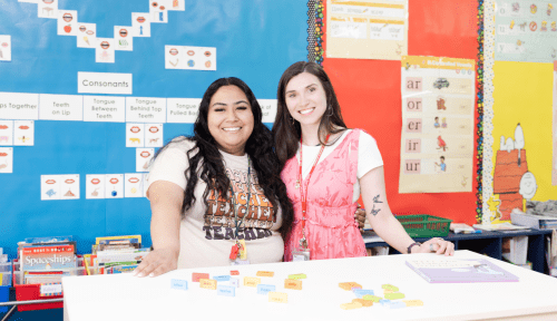 Two students stand in a classroom with education paraphernalia around them