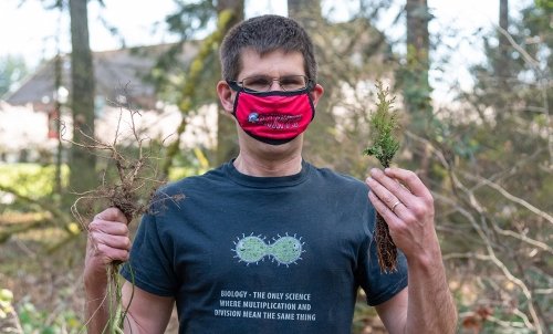 Dr. Bode, donning a Saint Martin's facemask, holds up two saplings outdoors at a sapling planting.