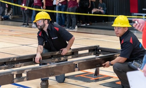 Two students in yellow hardhats working with a big metal device a caution tape-lined gymnasium, an audience looking on.