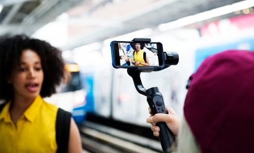 A young woman speaks in front of subway tracks while being filmed a cell phone being held in a steadying device.