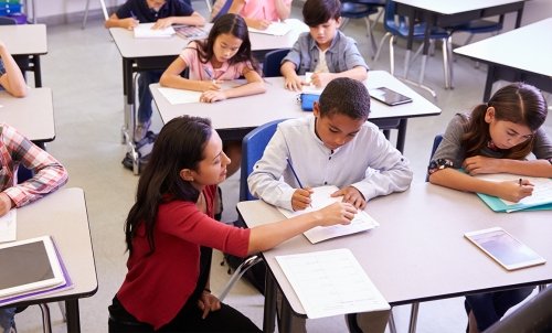 In a room full of children sharing desks, a teacher kneels at a desk to assistant a student doing some work.