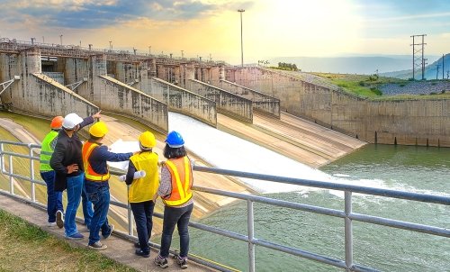 A group of people in hardhats and safety vests stand at a railing looking out a dam and the river.